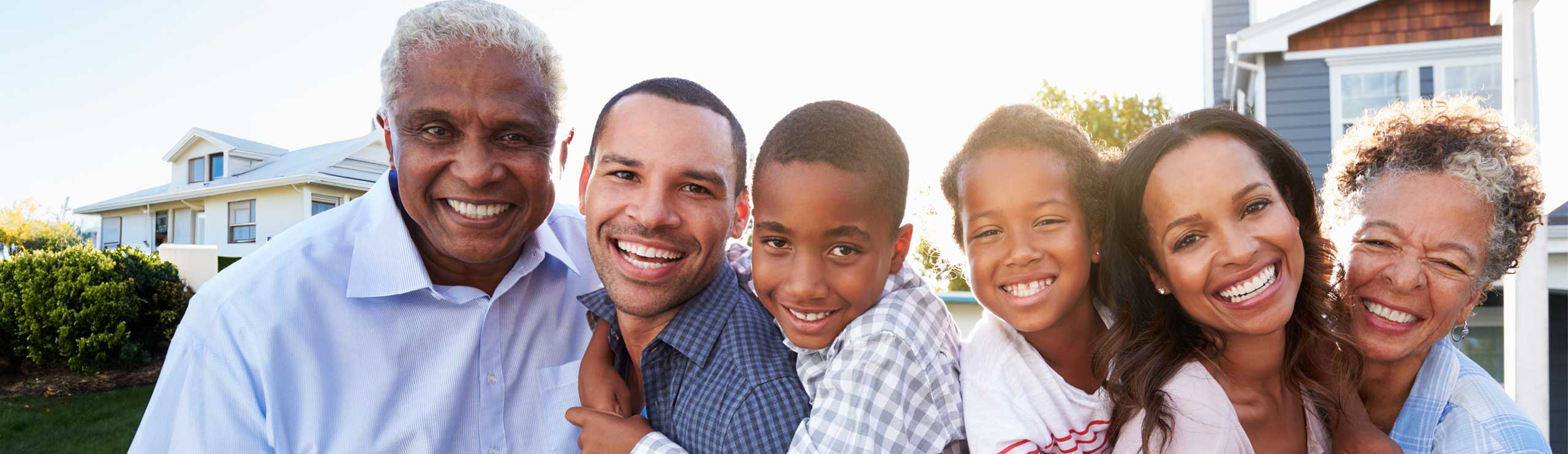 Three generations of a smiling family—grandparents, parents, and children—stand closely together in front of a suburban home, representing the supportive and inclusive nature of Indiana Medicaid Waiver caregiver services.