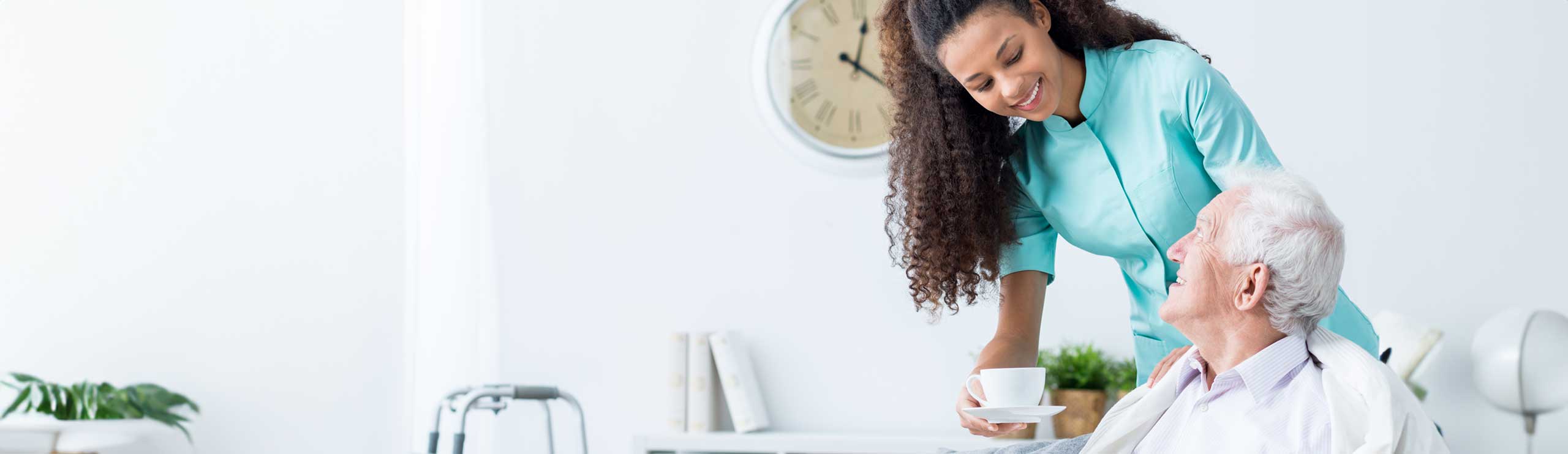 A smiling female caregiver in a teal uniform hands a cup of tea to a senior man sitting comfortably at home. The caregiver gently touches his shoulder, and they exchange warm smiles, reflecting trust, compassion, and attentive in-home care.