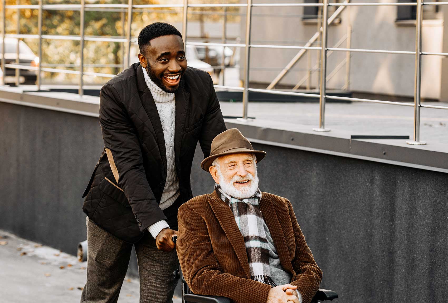 A cheerful young caregiver pushes a smiling senior man in a wheelchair outdoors on a sunny day, both dressed sharply and warmly, they share moment of joy. The scene reflects compassionate and engaging private-duty home care.